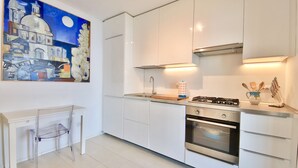 Una cocina moderna con gabinetes blancos, estufa a gas, horno y lavaplatos. En la pared, hay un cuadro de un paisaje urbano.
