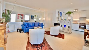 Una sala de estar moderna con un sofá azul, sillones blancos y una zona de cocina con armarios blancos y una mesa de comedor.