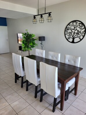 A dining area with a dark wooden table, six white upholstered chairs, a potted plant, a wall-mounted TV, and a decorative tree artwork.