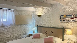 A bedroom with a stone wall, a bed with two pillows, a nightstand with a lamp, and a window with curtains. - Country house near SPILI with peaceful garden, orange trees and jacuzzi (Mixorouma)