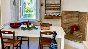 A dining area with a white table, two wooden chairs, a red bowl, a bottle of wine, and a basket of grapes. A window with curtains and a view of greenery is visible. - Country house near SPILI with peaceful garden, orange trees and jacuzzi (Mixorouma)
