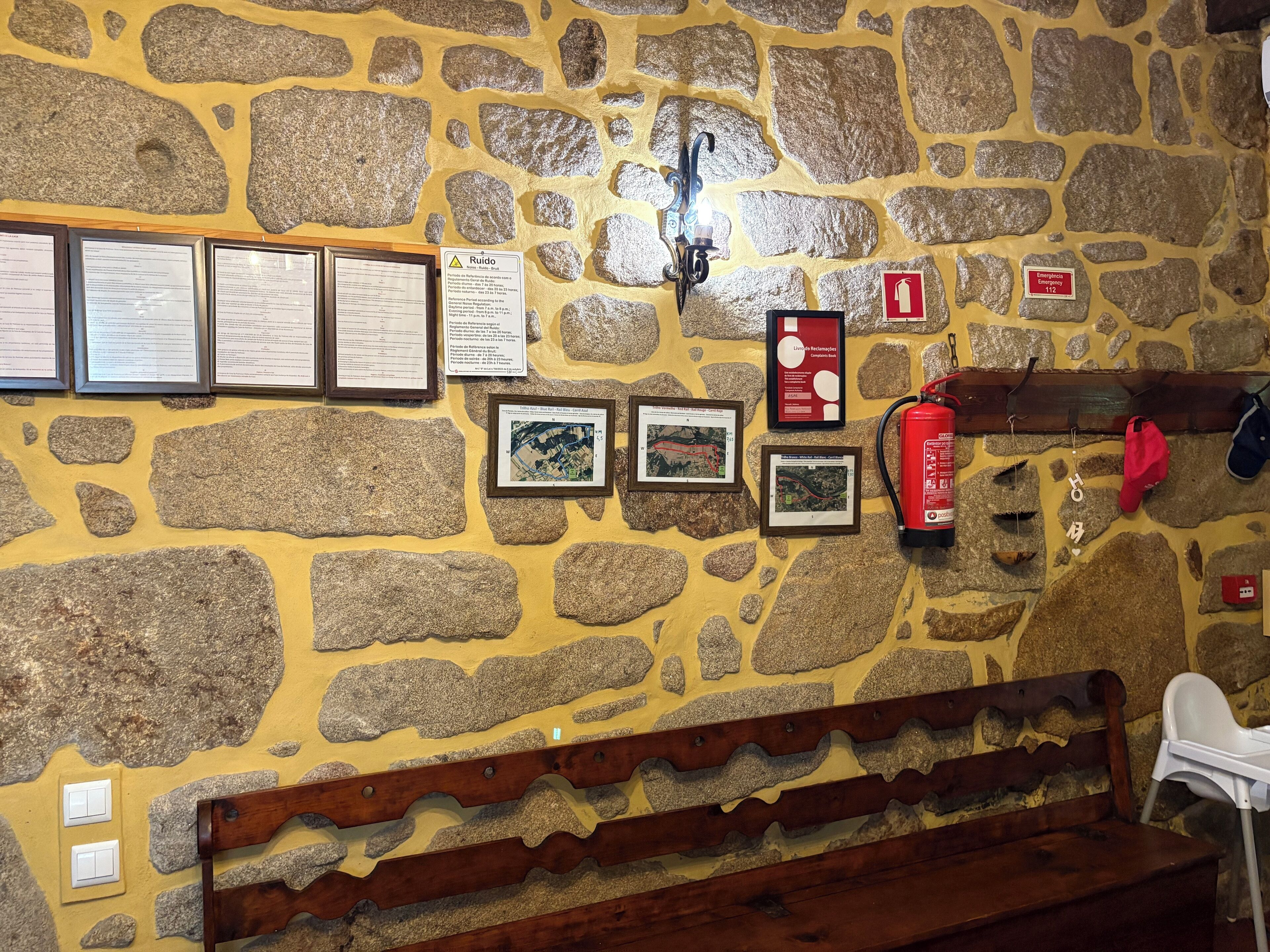 Een stenen muur met ingelijste documenten, een brandblusser en een houten bank.