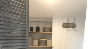 A hallway with wooden flooring, a grey wall, and a white ceiling. There are three shelves with various storage containers and a wall-mounted hook.