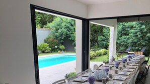 A long dining table set for a meal with a view of a swimming pool and greenery outside.