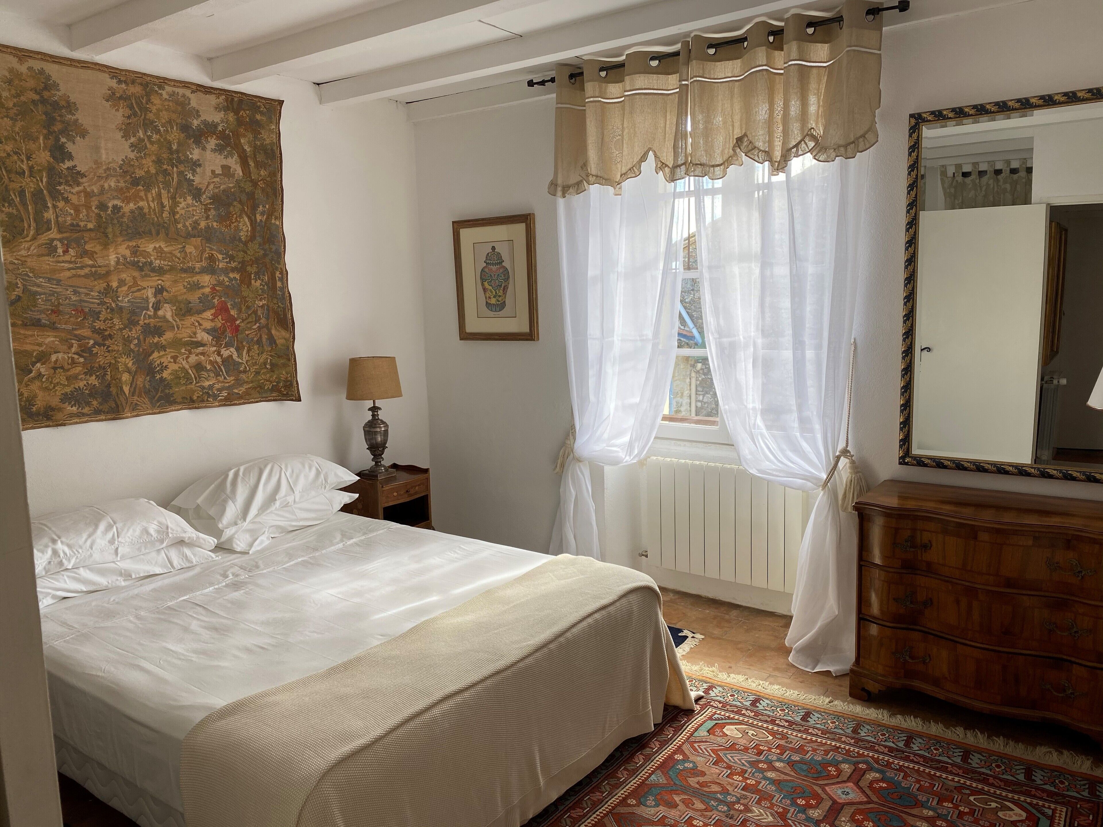 A bedroom with a bed, a nightstand, a lamp, a window with sheer curtains, a radiator, a dresser, and a large tapestry on the wall.