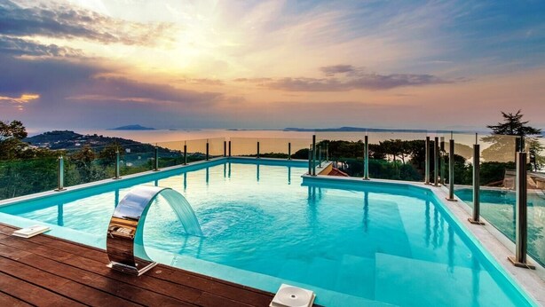 Ein rechteckiger Infinity-Pool mit klarem, blauem Wasser, umgeben von einer Holzterrasse und einem Glasgeländer, mit Blick auf eine malerische Landschaft mit Hügeln und einem Gewässer unter einem Sonnenuntergangshimmel.
