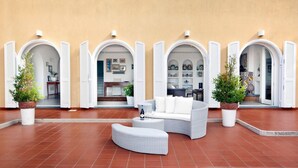 Ein Sitzbereich im Freien mit einer weißen Couch und Hocker, umgeben von weißen Fensterläden und Topfpflanzen.
