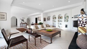 Ein geräumiges Wohnzimmer mit einer Couch, einem Couchtisch und Sesseln. Regale mit Dekorationselementen und einem Fernseher sind ebenfalls vorhanden.