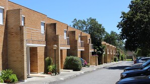 Une rangée d’immeubles à appartements en briques, dotés de balcons, avec un trottoir pavé et des voitures garées.