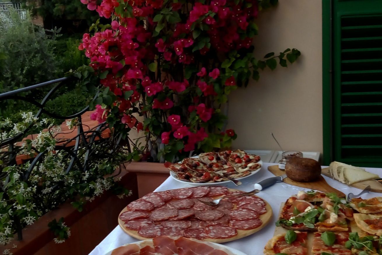 Une table dressée avec divers aliments, notamment des viandes en tranches et des pizzas, sur un balcon offrant une vue sur la verdure et une plante à fleurs rouges.