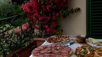 Une table dressée avec divers aliments, notamment des viandes en tranches et des pizzas, sur un balcon offrant une vue sur la verdure et une plante à fleurs rouges.