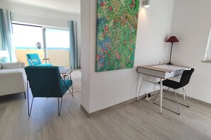 A modern hotel room with a balcony, a desk, a chair, and a large floral painting on the wall.
