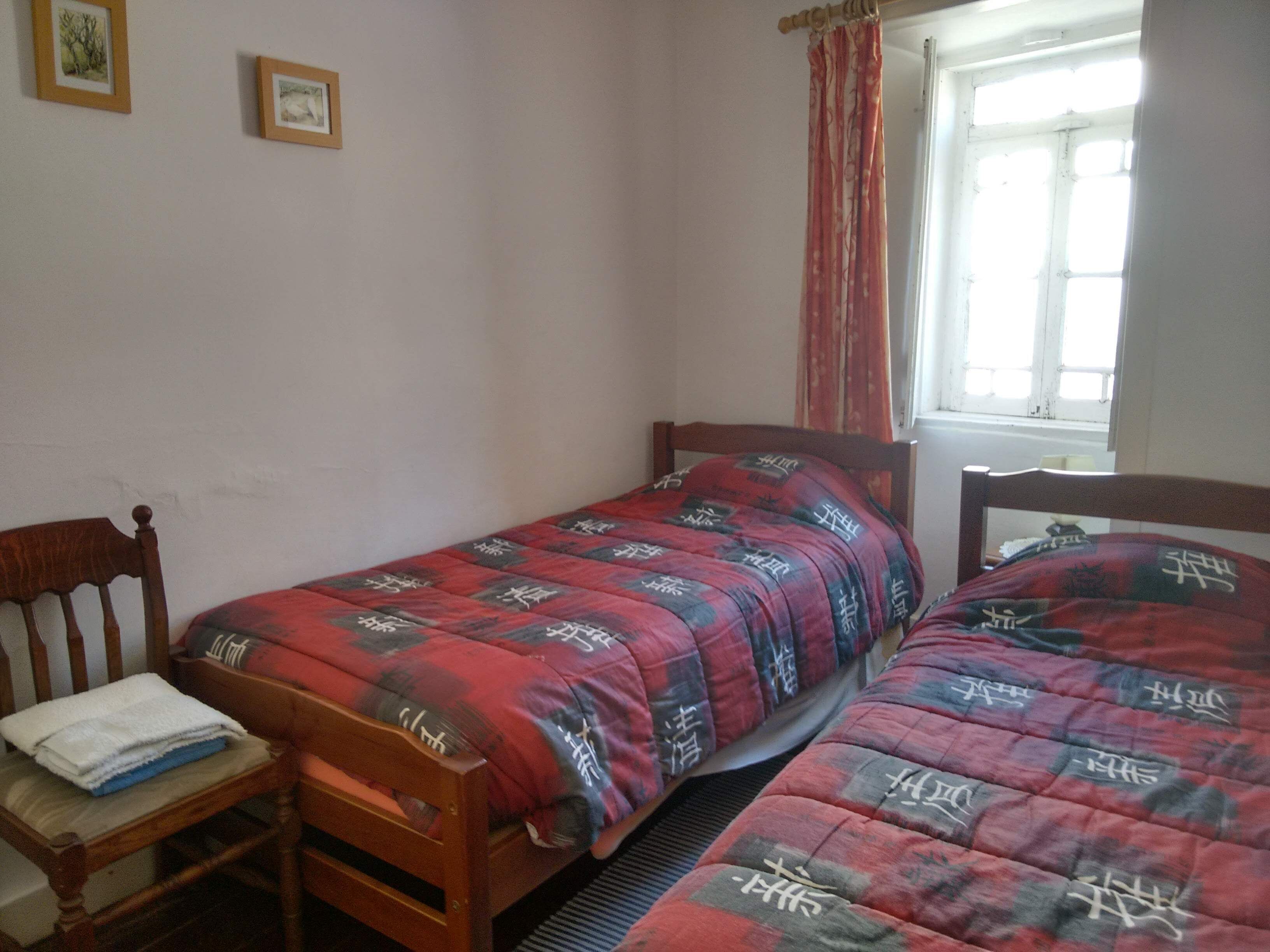A bedroom with two single beds, a wooden chair, a window with curtains, and two framed pictures on the wall.