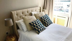 Una cama con cabecera capitonada, dos almohadas estampadas, una colcha blanca, una lámpara de noche y vista a un puerto deportivo a través de una ventana.