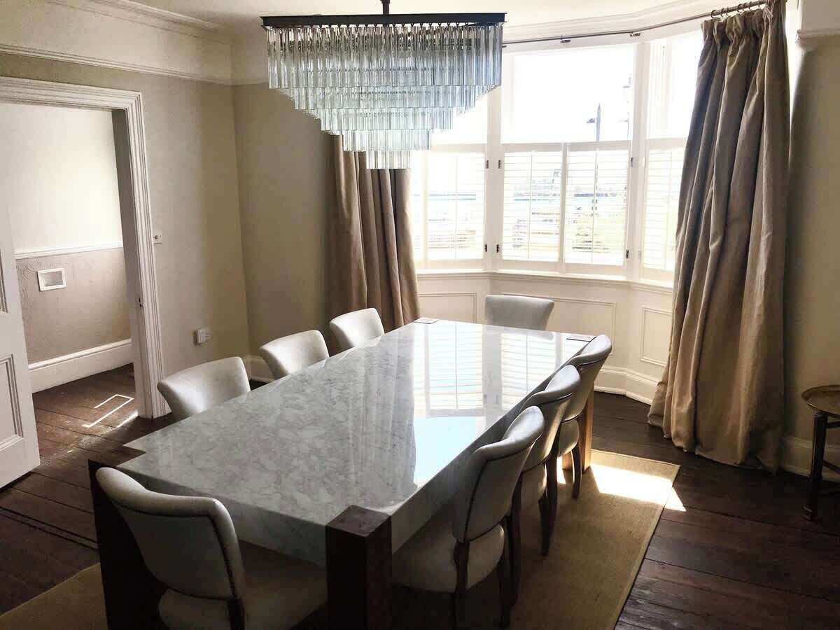 Comedor con una mesa de mármol, seis sillas, una araña y ventanas grandes con cortinas.