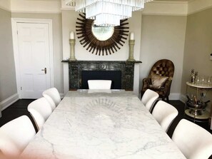 A dining room with a marble table, white chairs, a fireplace, and a chandelier.