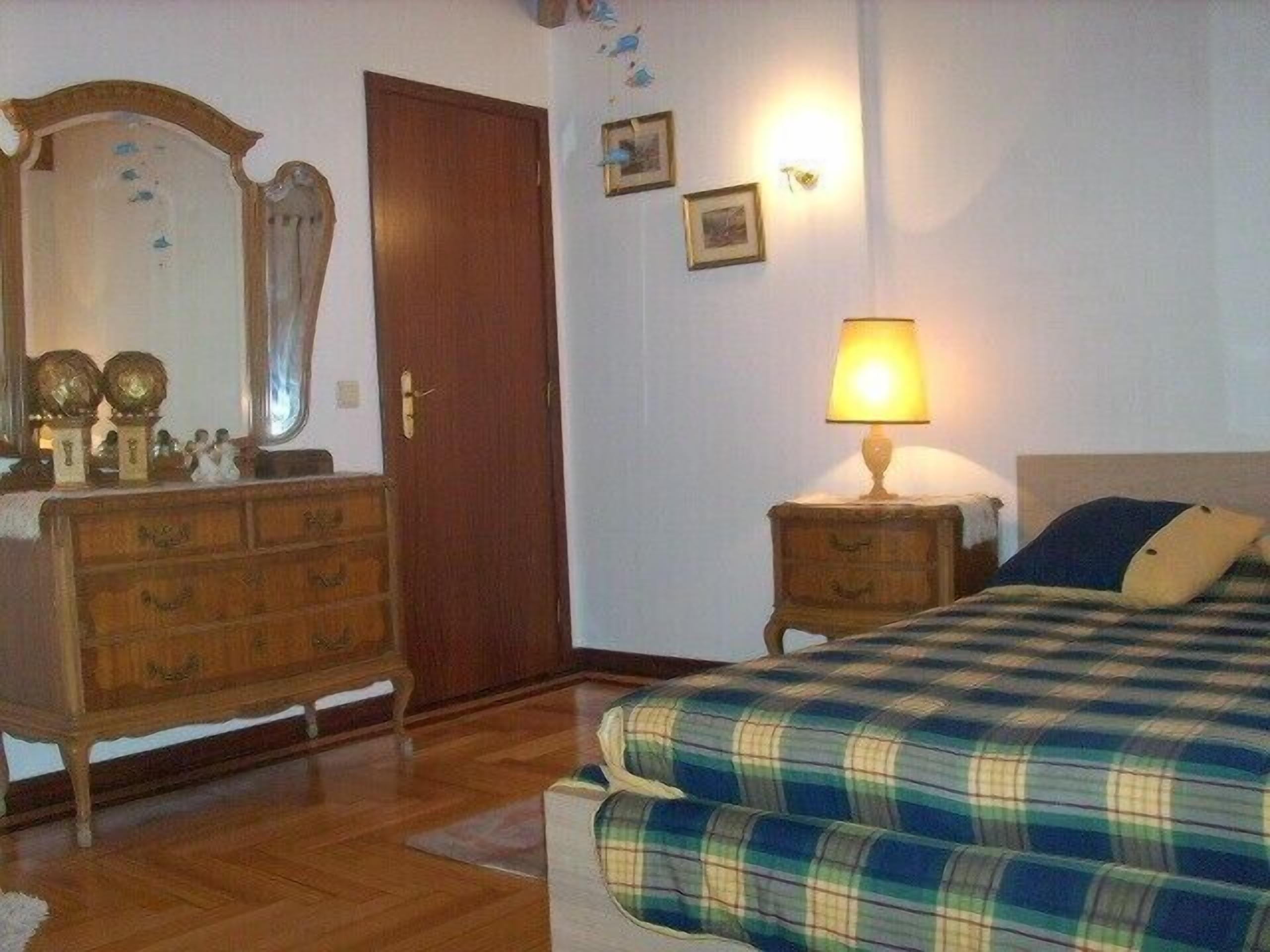 A bedroom with a wooden dresser, a bed with a plaid duvet, a nightstand with a lamp, and a door.