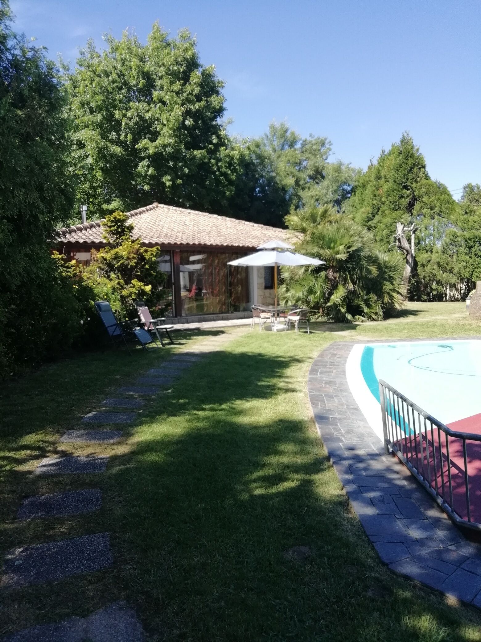 A backyard with a swimming pool, a patio with chairs and an umbrella, and a house with a tiled roof.