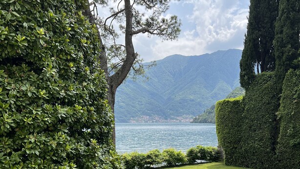 Eine hölzerne Bank auf einem Balkon mit Blick auf einen gepflegten Rasen, einen See und Berge.