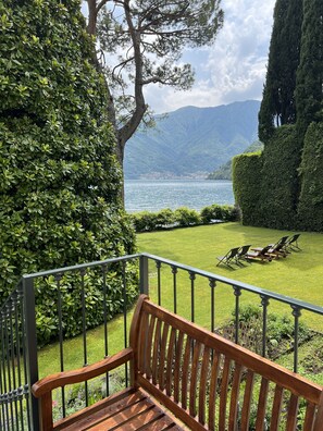 A wooden bench on a balcony overlooking a well-manicured lawn with a view of a lake and mountains.