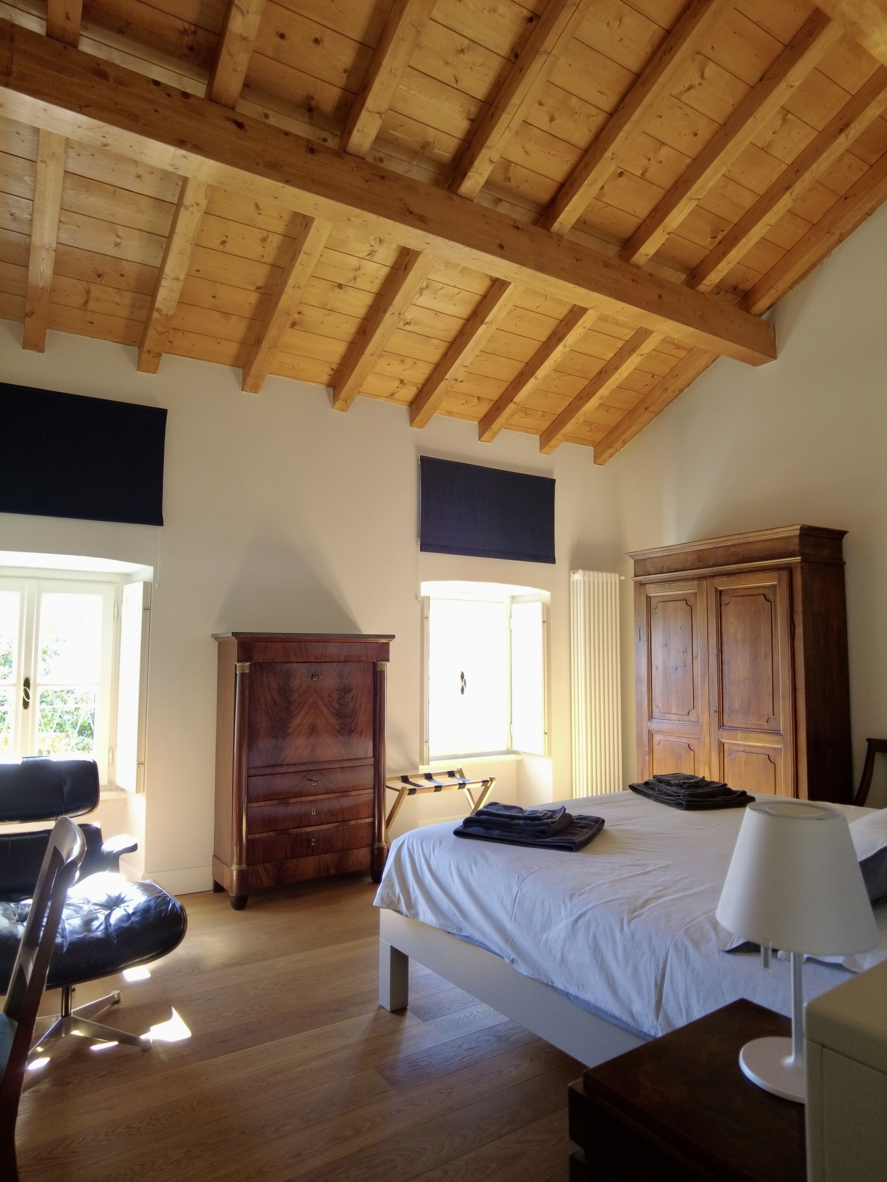 A bedroom with a wooden ceiling, a bed with white linens, a wooden dresser, a chair, and a desk.