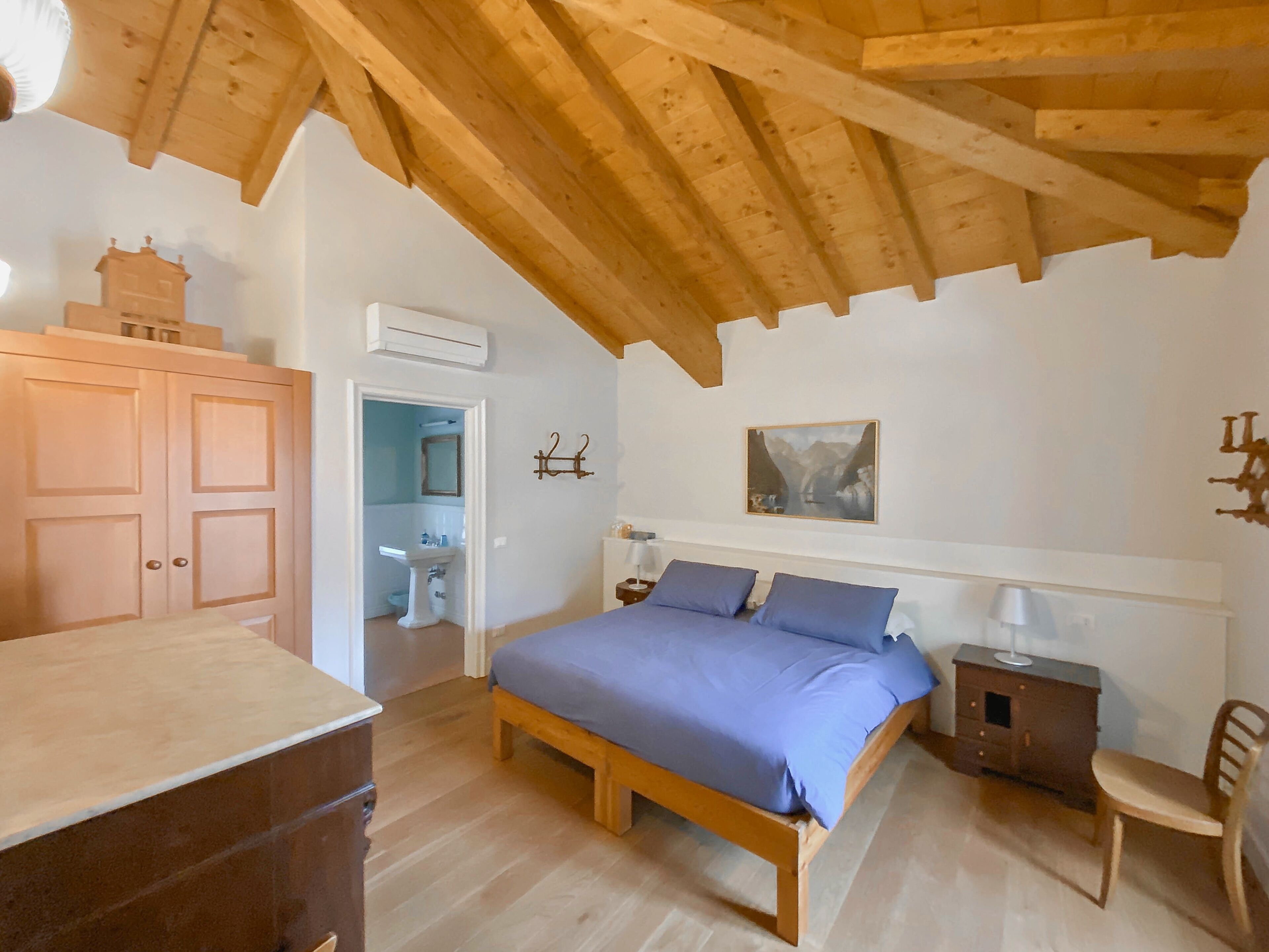 Een slaapkamer met een houten bed, een houten dressoir, een bureau en een stoel. Een badkamer is zichtbaar door een geopende deur.