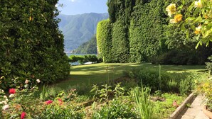 Ein gepflegter Garten mit einer Vielfalt an Pflanzen und Blumen, einem ordentlich geschnittenen Heckenstreifen und Blick auf einen See mit Bergen im Hintergrund.