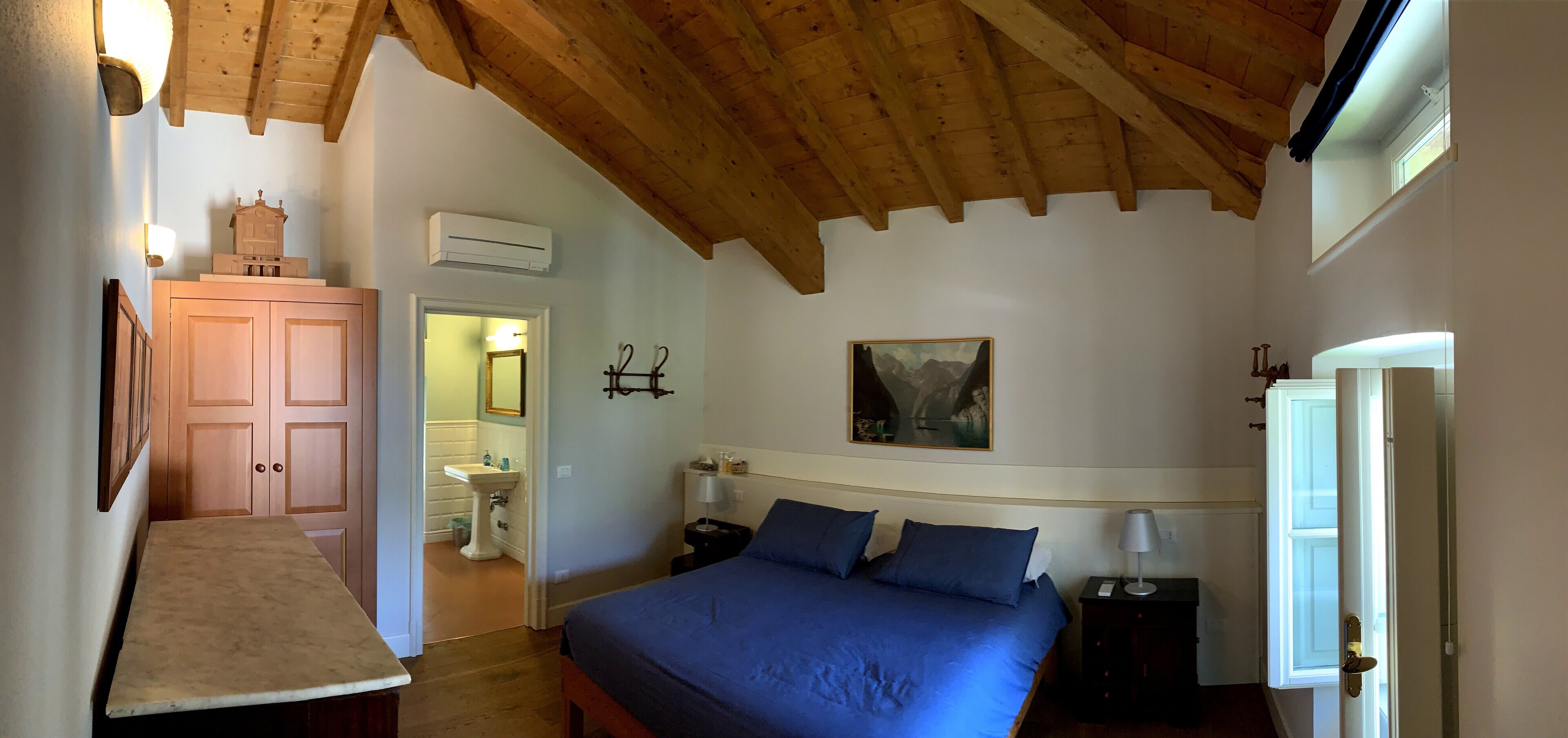 A bedroom with a wooden ceiling, a bed with blue bedding, a nightstand, a wall-mounted lamp, and a painting above the bed.