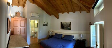 A bedroom with a wooden ceiling, a bed with blue bedding, a nightstand, a wall-mounted lamp, and a painting above the bed.