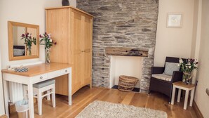 A room with a wooden desk, a mirror, a stone wall, a wicker chair, and a small table with flowers.