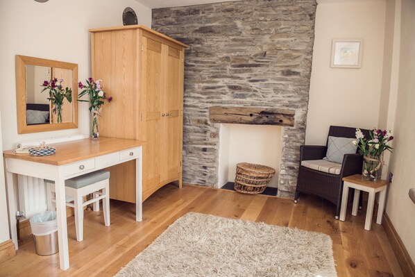 A room with a wooden desk, a mirror, a stone wall, a wicker chair, and a small table with flowers.
