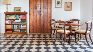 Een kamer met een geruite vloer, een houten boekenkast, een houten deur, een eettafel met stoelen en ingelijste foto’s aan de muur.