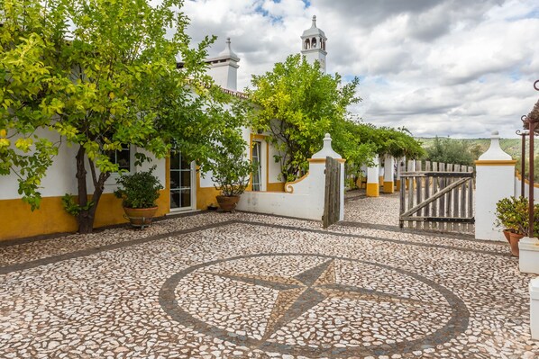 A courtyard with a tiled floor, potted plants, a gate, and a building with a bell tower.