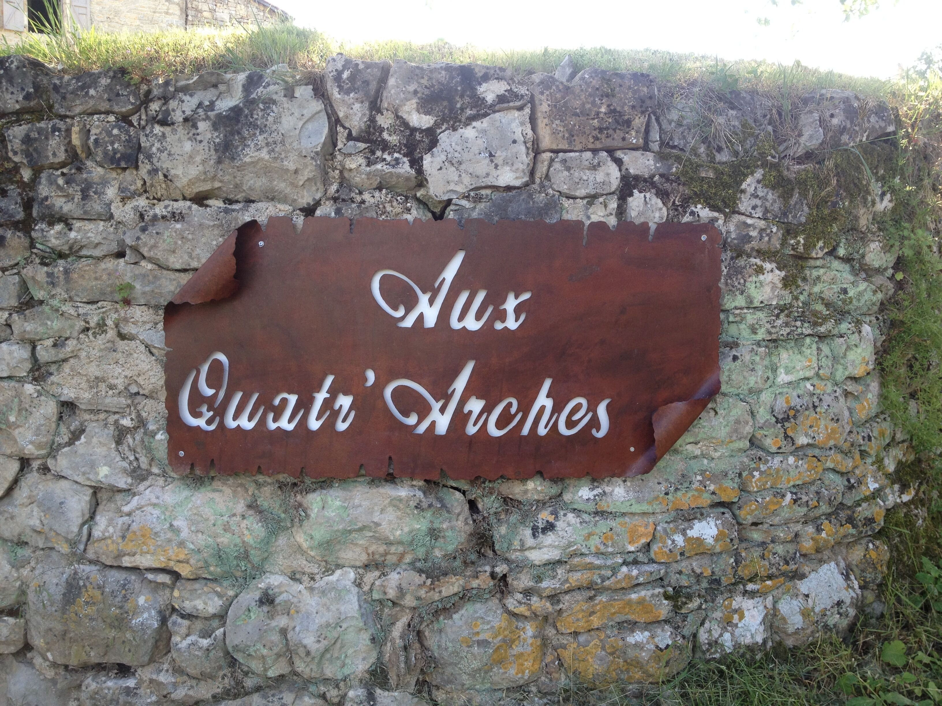 Ein verwittertes Metallschild mit der Aufschrift 'Aux Quatre Arches' an einer Steinmauer.