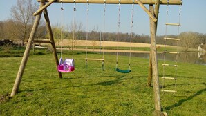 Een houten schommel met twee schommels, één roze en één groen, in een parkachtig gebied met gras en een waterpartij, met bomen op de achtergrond.
