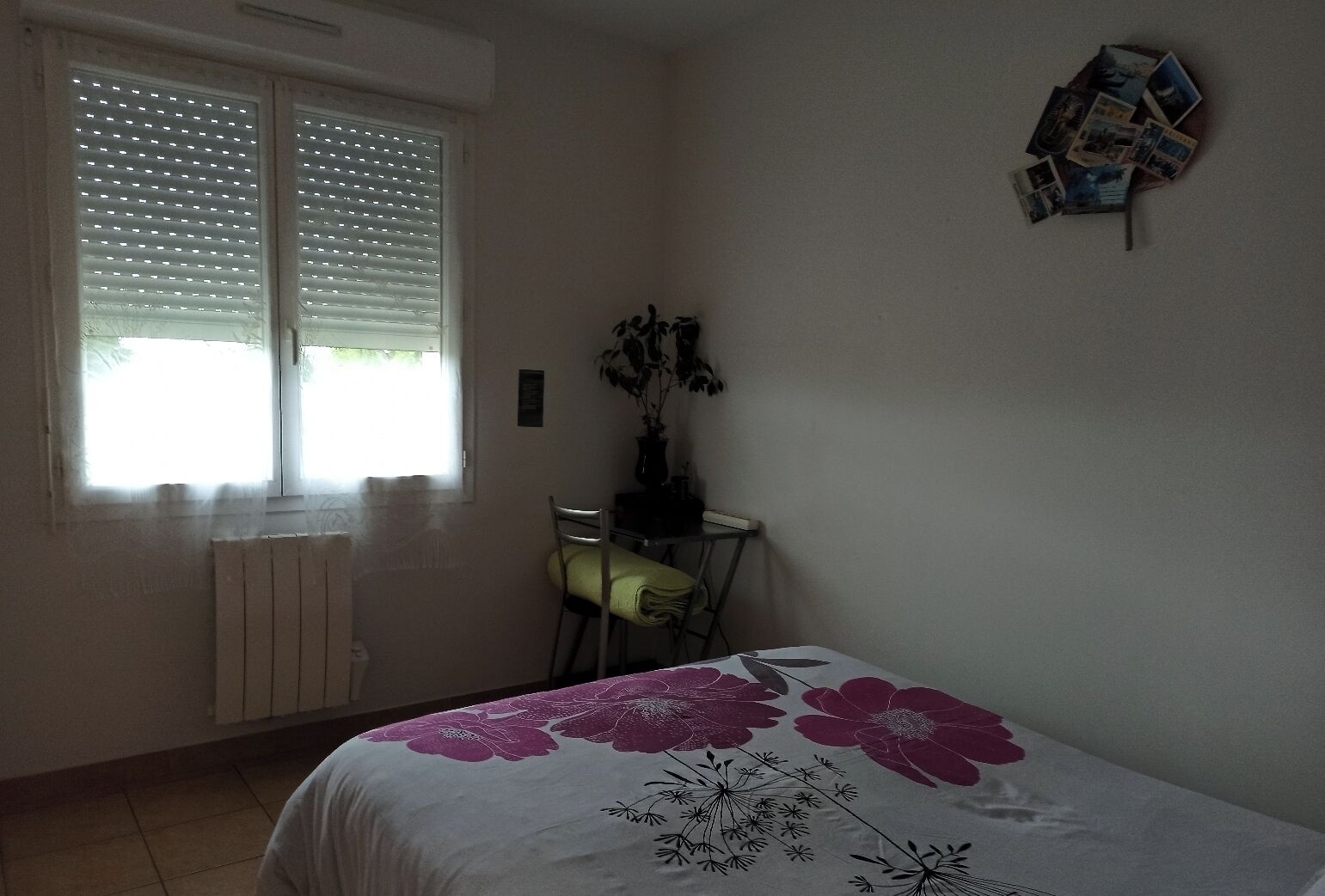 Une chambre à coucher comprenant un lit, une chaise, un radiateur, une fenêtre avec des stores et une étagère fixée au mur où sont posées des photos.