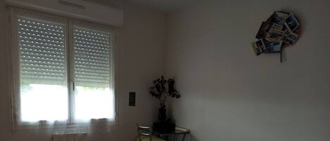 Une chambre à coucher comprenant un lit, une chaise, un radiateur, une fenêtre avec des stores et une étagère fixée au mur où sont posées des photos.