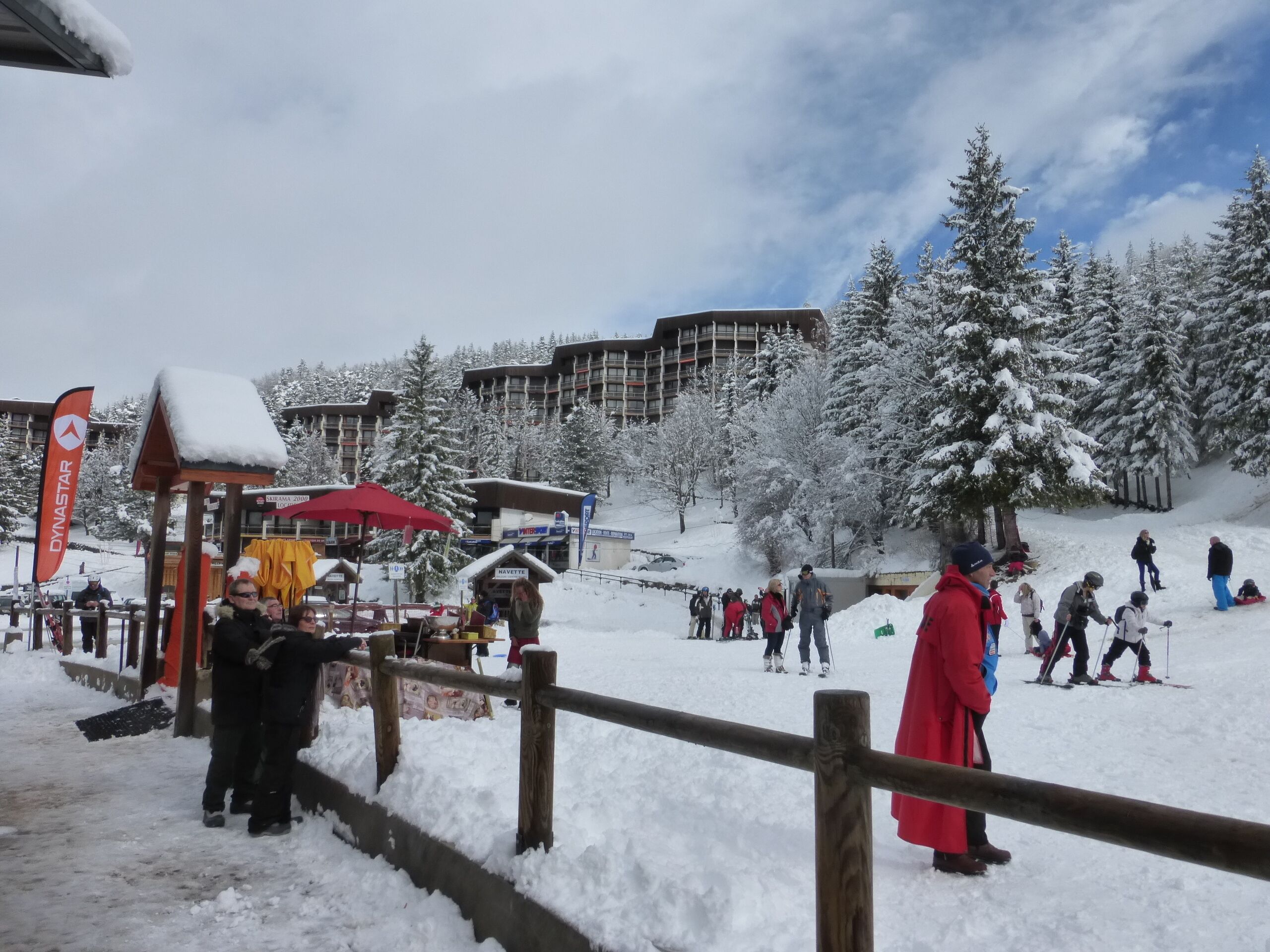 Un espace extérieur enneigé, avec une clôture en bois, un parasol rouge et des personnes qui font du ski.