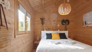 A wooden cabin bedroom with a bed, a hanging dreamcatcher, a wall-mounted mirror, and a window with a view of greenery. - The Sea Lodge - Free Parking (Zandvoort)