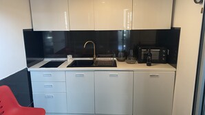 Cocina compacta con armarios blancos, salpicadero negro, un lavamanos con grifo dorado, microondas y una silla roja.