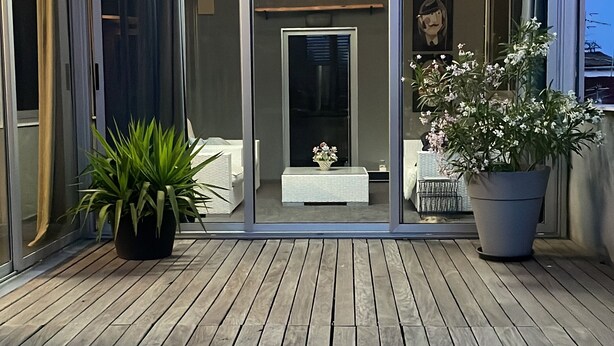 Una terraza de madera con plantas en macetas, un acceso por puerta de cristal y vistas a una habitación con cama y mesa.