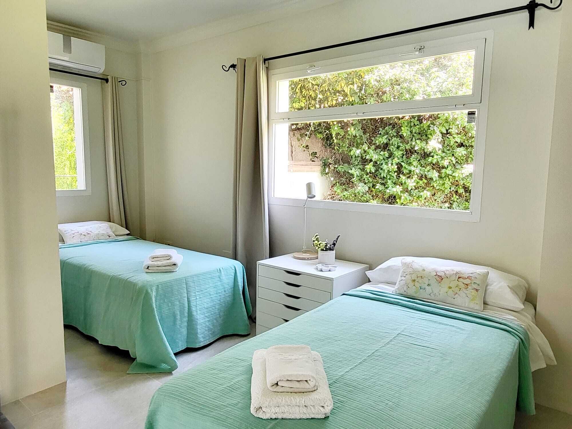 A bedroom with two beds, a small bedside table, a window with a view of greenery, and a ceiling fan.