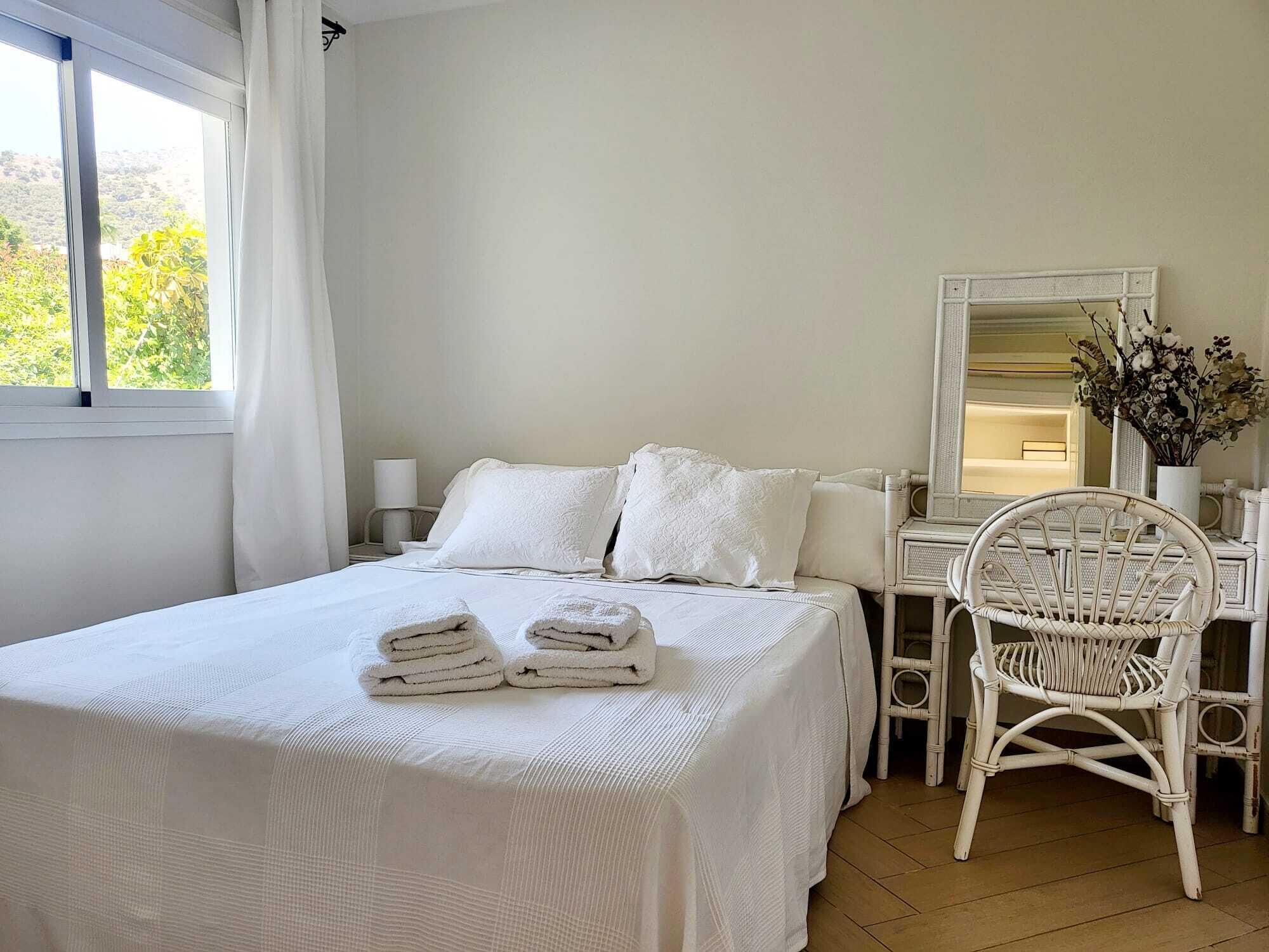 Een slaapkamer met een bed, witte kussens, een stoel, een spiegel en een raam met gordijnen.