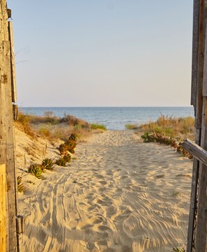 Ein sandiger Küstenweg führt zum Meer, umgeben von Holzbauten und Vegetation.