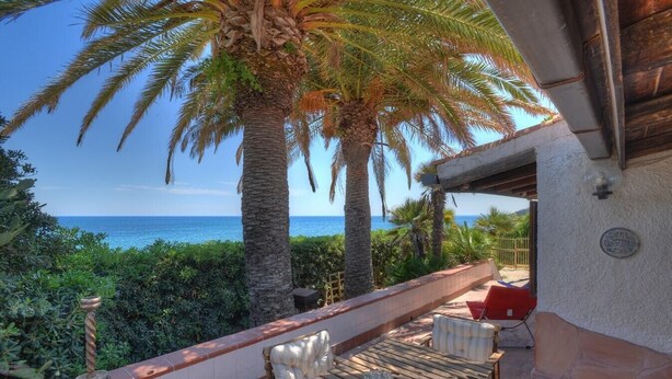 Eine Terrasse mit Holztisch und -stühlen mit Blick auf einen Strand, der von Palmen und dem Meer gesäumt wird.