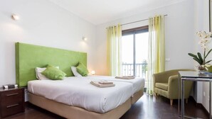 Habitació d'hotel amb un llit gran, un reposacapses verd, una taula de treball amb una cadira i una finestra amb cortines.