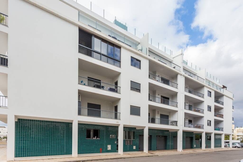 A multi-story residential building with balconies, a tiled ground floor, and a clear sky.