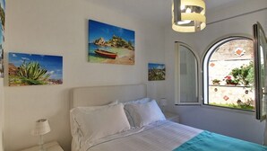 A bedroom with a bed, a nightstand, a lamp, a window with a view of a garden, and wall art.