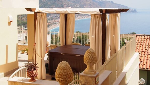 A balcony with a wooden table and chairs, a potted plant, and a view of the sea and mountains.