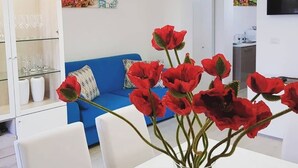 A modern living room with a blue sofa, a white dining table with a vase of red flowers, and a kitchen area in the background.
